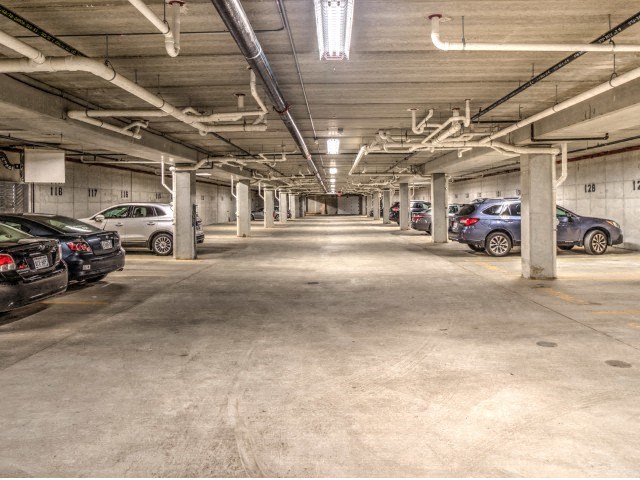 Dimly lit underground parking garage with concrete pillars and cars parked on both sides. The space appears orderly and empty, conveying a quiet atmosphere at Stitchweld Apartments.