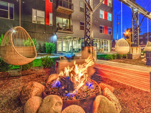 A cozy outdoor fire pit surrounded by stones, set in a modern apartment courtyard with colorful lighting and hanging chairs, creating a relaxed ambiance at Stitchweld Apartments.