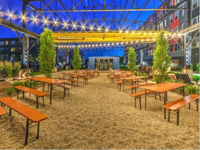 Outdoor garden at dusk with empty wooden tables and benches on gravel. Strings of lights hang above, creating a cozy, inviting atmosphere at Stitchweld Apartments.