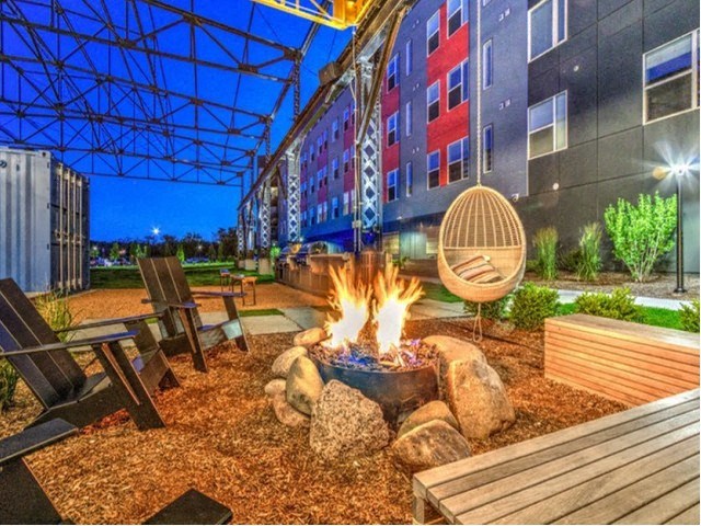 Vibrant outdoor patio with a crackling fire pit surrounded by chairs. A modern building with colorful lights at Stitchweld Apartments.