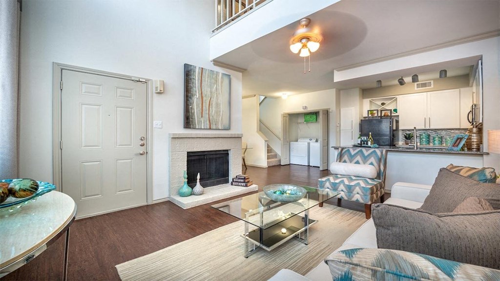 Bright living room here at Lakewood on the Trail Apartment Homes with high ceilings, featuring a beige sofa, glass coffee table, fireplace, abstract art, and modern kitchen. Airy and inviting ambiance.