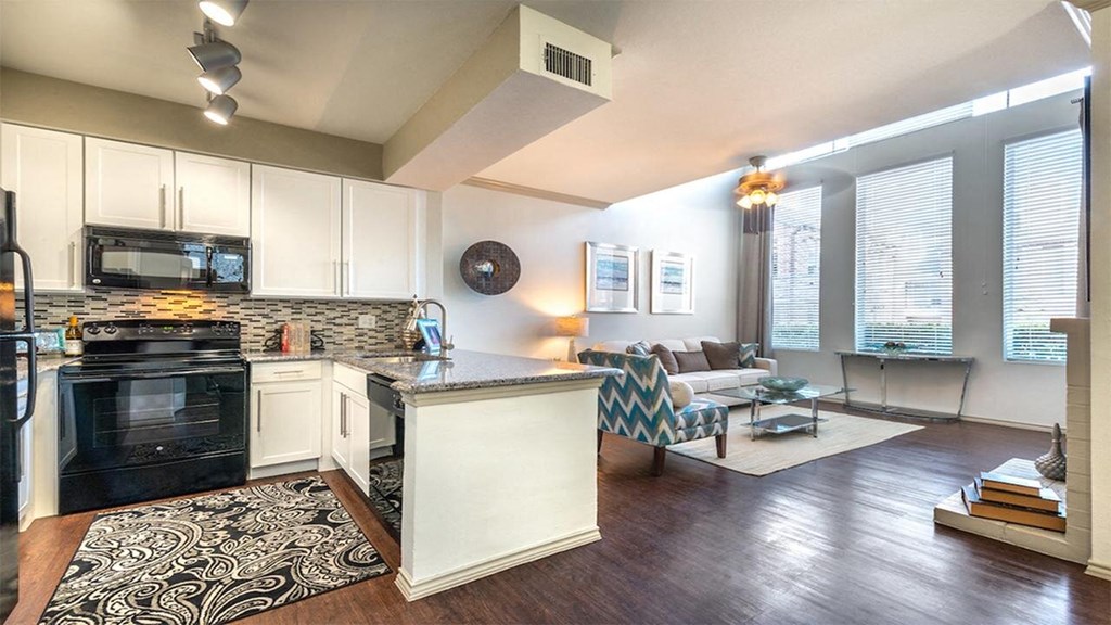Modern open-plan kitchen and living area here at Lakewood on the Trail Apartment Homes with wood floors. White cabinets, black appliances, and a patterned rug accent the kitchen. The living space includes a cozy sofa, patterned armchair, and tall windows for natural light.