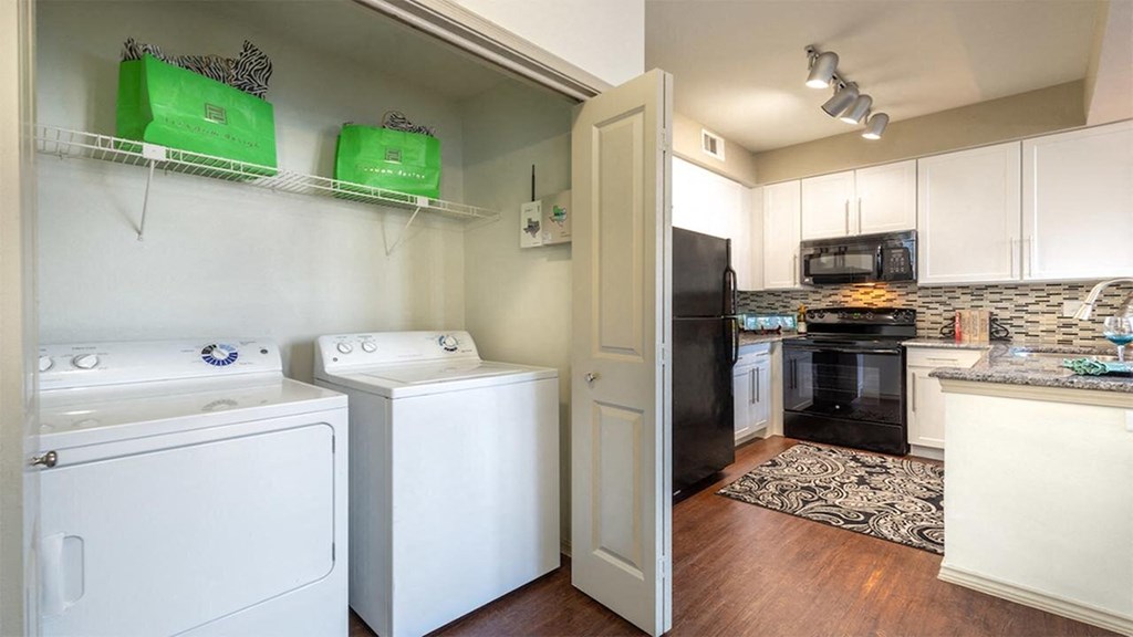Laundry area here at Lakewood on the Trail Apartment Homes with a white washer and dryer beside a modern kitchen. The kitchen features white cabinets, a black fridge, and an ornate rug.