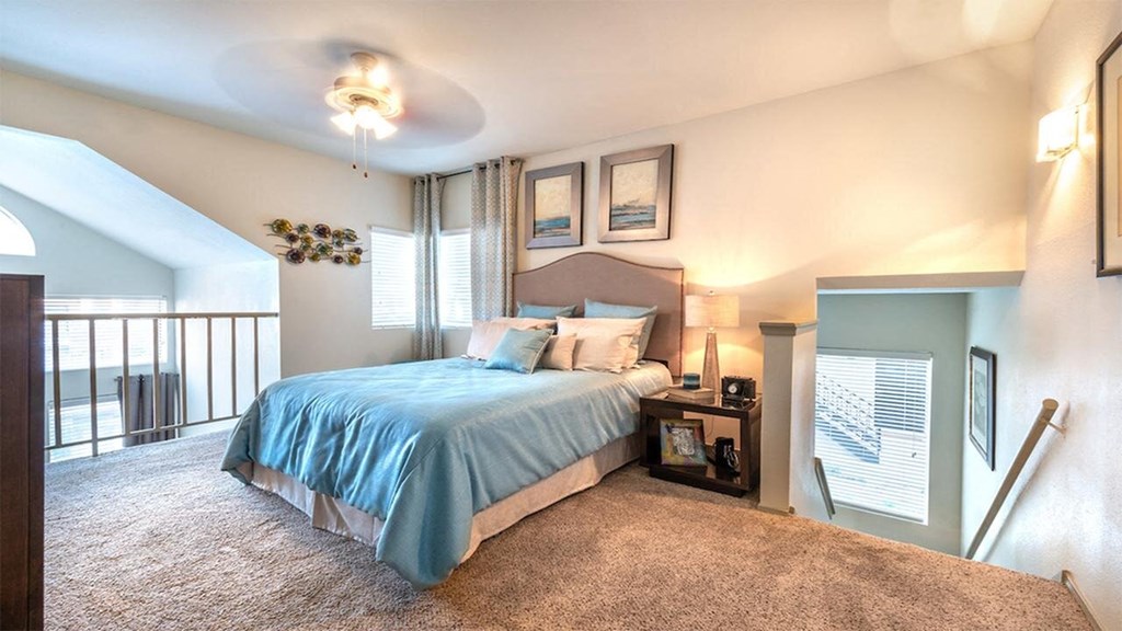 A cozy bedroom here at Lakewood on the Trail Apartment Homes with soft beige carpet features a bed with blue bedding, flanked by a bedside table and lamp. Sunlight filters through two windows, enhancing the serene, airy ambiance.