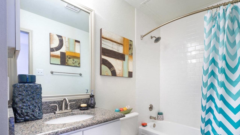 Modern bathroom here at Lakewood on the Trail Apartment Homes with granite countertop, abstract art, and a teal chevron shower curtain. Bright and clean with a serene, stylish atmosphere.