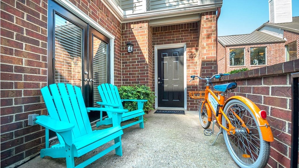 A cozy brick patio here at Lakewood on the Trail Apartment Homes with two bright turquoise chairs beside a glass door, an orange bicycle leaned against a fence, and a black front door exuding a cheerful, welcoming vibe.