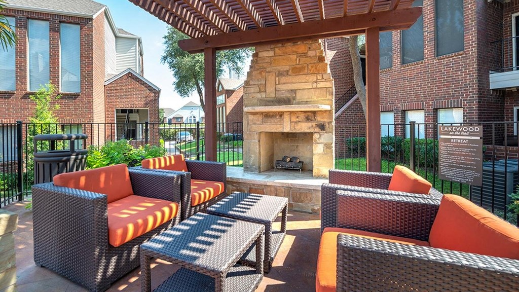 Outdoor patio here at Lakewood on the Trail Apartment Homes with wicker furniture and orange cushions surrounding a stone fireplace under a wooden pergola. Brick buildings and greenery in the background.
