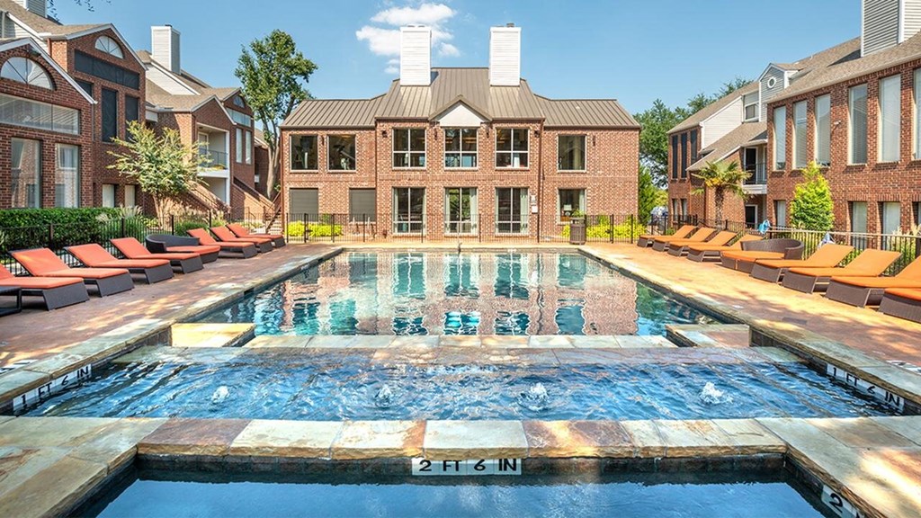 Courtyard here at Lakewood on the Trail Apartment Homes with a large outdoor pool and jacuzzi in the foreground, surrounded by orange lounge chairs, brick buildings, and trees under a clear blue sky.