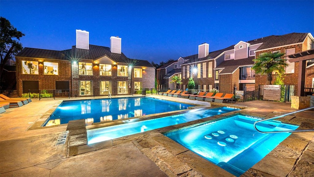 Outdoor pool area here at Lakewood on the Trail Apartment Homes at dusk with glowing lights. Brick buildings surround the pool; lounge chairs are neatly arranged. Palm trees add a relaxing touch.