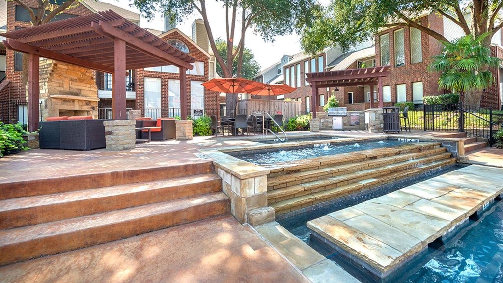 Outdoor pool area here at Lakewood on the Trail Apartment Homes with a jacuzzi, surrounded by wooden pergolas, patio seating, and red umbrellas. There are trees, brick buildings, and a relaxed ambiance.