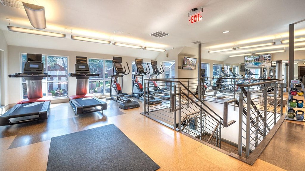 Bright gym interior here at Lakewood on the Trail Apartment Homes with treadmills and ellipticals near large windows. A staircase leads down, and colorful kettlebells are visible, creating an energetic atmosphere.
