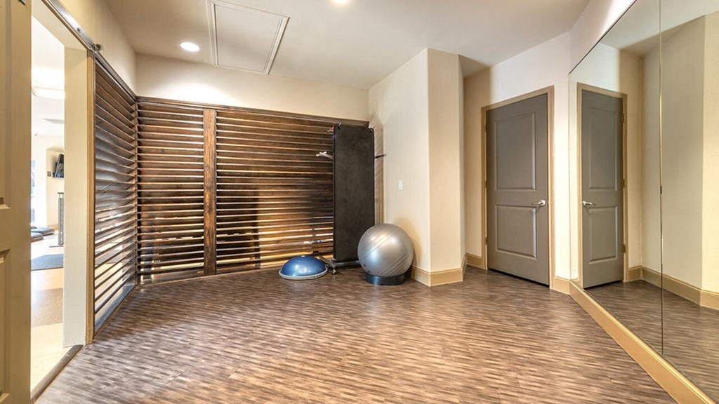 A small, modern home gym here at Lakewood on the Trail Apartment Homes with light wood flooring, exercise ball, balance trainer, and a large mirror on the right wall. The space is well-lit and inviting.