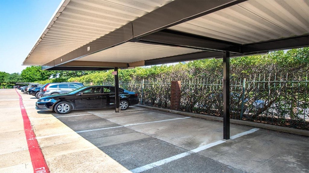 Covered parking area here at Lakewood on the Trail Apartment Homes with several cars parked in marked spaces. Metal roof structure provides shade. Fenced greenery in the background; calm, sunny day.