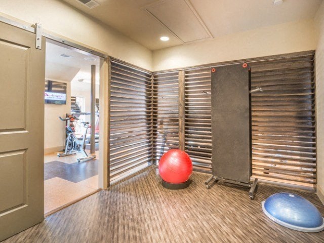 Small gym room here at Lakewood on the Trail Apartment Homes with wooden walls, featuring a red exercise ball, blue BOSU ball, and a foldable wall-mounted bench. Open door leads to a larger gym space.