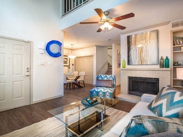 Modern living room here at Lakewood on the Trail Apartment Homes with a white couch and chevron pillows, glass coffee table, and a fireplace. Dining area and stairs are visible in the background.