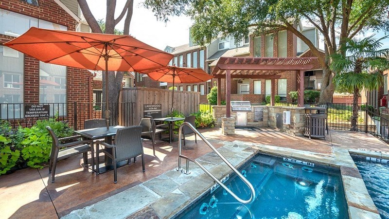 Outdoor patio here at Lakewood on the Trail Apartment Homes with tables and orange umbrellas beside a blue hot tub. A pergola with a grill is in the background, surrounded by lush greenery. Relaxed setting.