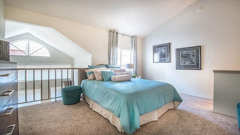 Spacious bedroom here at Lakewood on the Trail Apartment Homes with soft lighting, featuring a blue bedspread and matching pillows. Two framed artworks hang on a beige wall, exuding a calm ambiance.