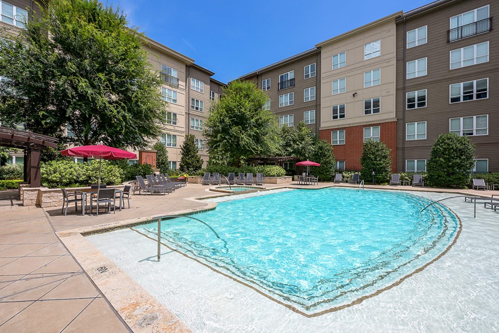 Cityplace Heights Apartment Homes courtyard with a large, inviting swimming pool. Surrounding the pool are lounge chairs and red umbrellas, set against a backdrop of leafy trees and a multi-story building under a clear blue sky.