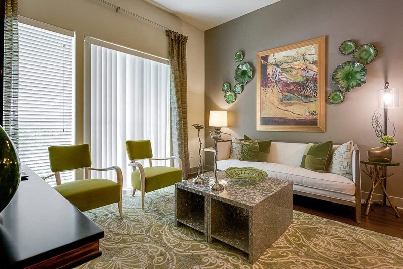 A stylish living room here at Cityplace Heights Apartment Homes with green accents. Two green chairs and a patterned rug face a sofa. Wall art includes abstract paintings and decorative plates. A lamp emits a warm glow.