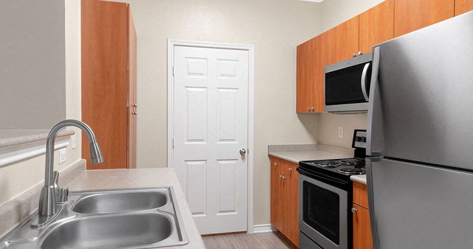 Modern kitchen here at Landing at Mansfield Apartment Homes with light wood cabinets, stainless steel appliances, and a double sink. A closed white door is centered in the back.