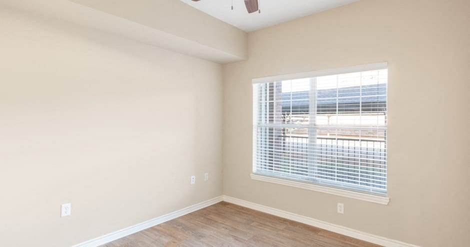 Empty bedroom here at Landing at Mansfield Apartment Homes with light gray walls, wooden floor, and a ceiling fan with light. A large window with blinds lets in sunlight, creating a bright atmosphere.