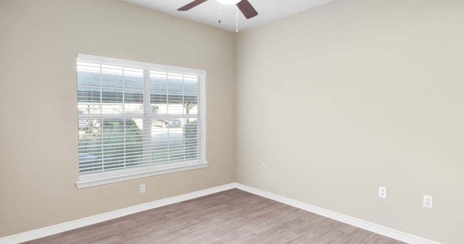 An empty bedroom here at Landing at Mansfield Apartment Homes with beige walls, a large window with blinds, light wooden floors, and a ceiling fan. The room appears bright and minimalist.