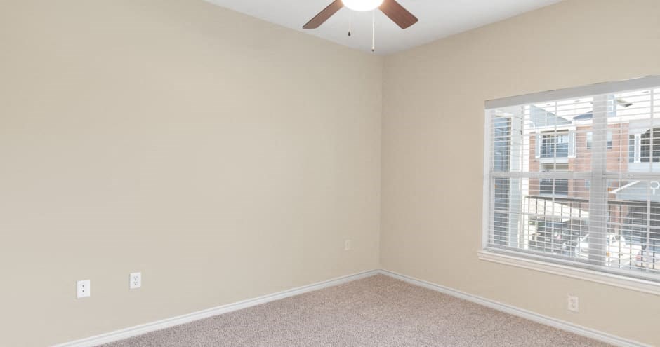 Empty bedroom here at Landing at Mansfield Apartment Homes with beige walls, carpeted floor, and ceiling fan. A large window with blinds lets in natural light, overlooking an exterior building.