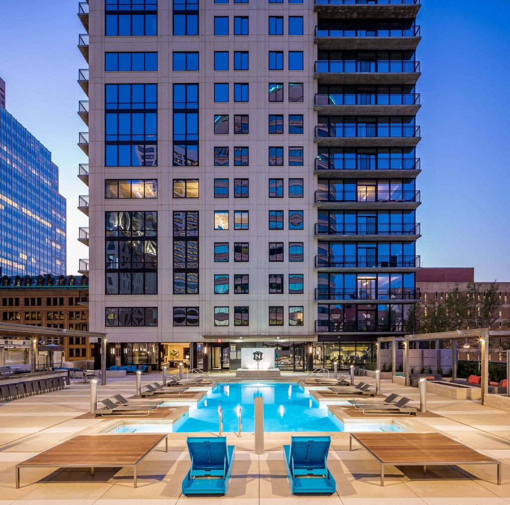 Rooftop pool area here at Nic on 5th Apartment Homes at dusk with modern high-rise backdrop. Two vibrant blue lounge chairs face the illuminated pool, creating a relaxed, luxurious ambiance.