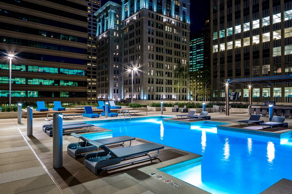 Rooftop pool here at Nic on 5th Apartment Homes, at night, surrounded by modern high-rise buildings with bright windows. Blue loungers and soft lighting create a relaxing, urban atmosphere.