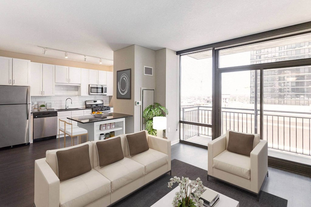 Modern open-plan living room and kitchen here at Nic on 5th Apartment Homes, featuring light beige sofas with brown cushions, a coffee table with flowers, and large windows leading to a balcony.