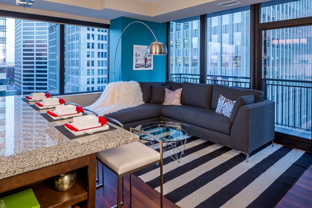 Modern living room here at Nic on 5th Apartment Homes with a corner grey sofa, blue accent wall, and a striped rug. Large windows reveal a cityscape. A table with dining settings sits nearby.
