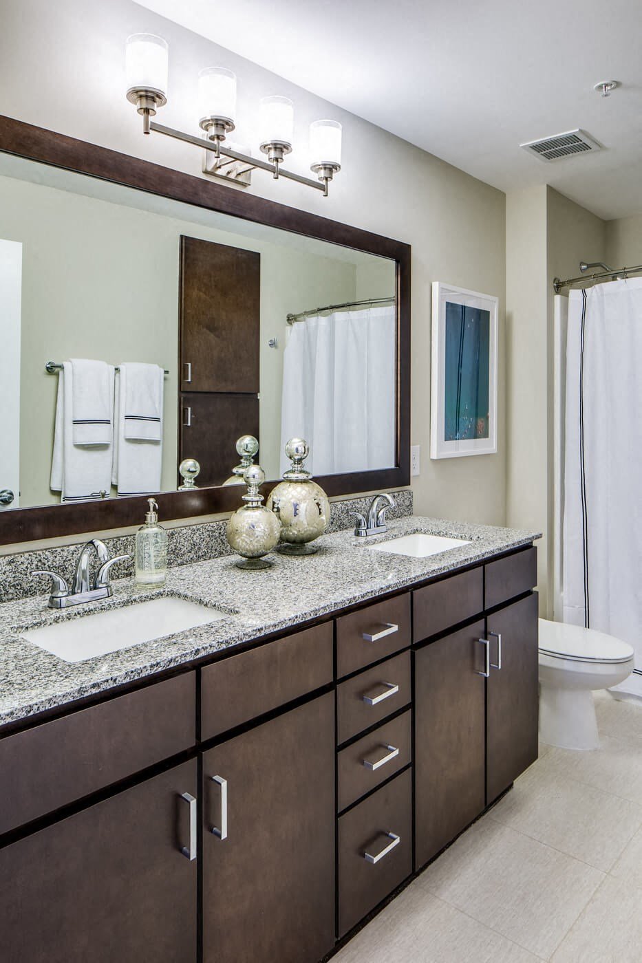 Modern bathroom here at Nic on 5th Apartment Homes with a granite double sink countertop, dark wood cabinets, and a large mirror. Bright lighting, decorative vases, and white towels add elegance.