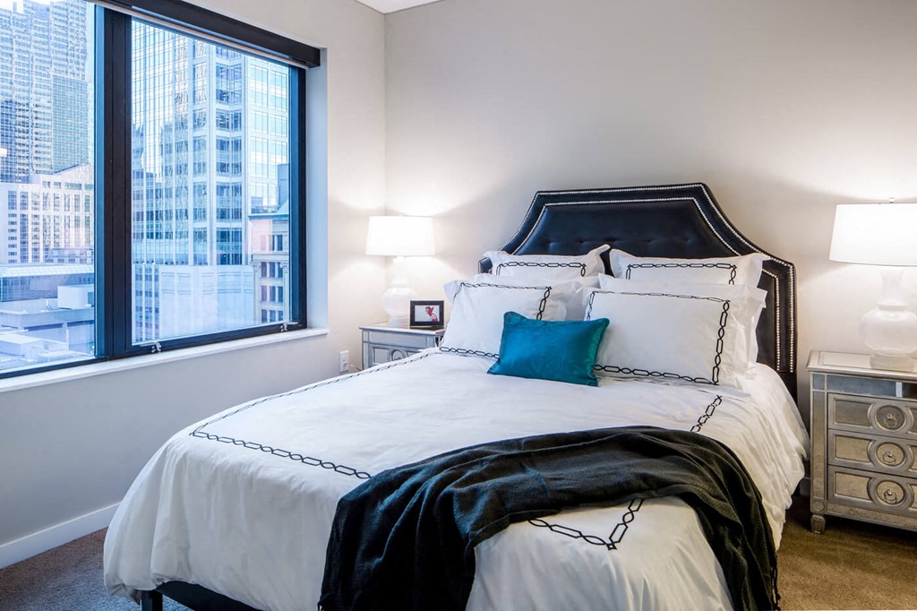 Modern bedroom here at Nic on 5th Apartment Homes with a dark headboard and white bedding accented by a teal pillow and dark throw. Large window shows cityscape buildings. Calm ambiance.