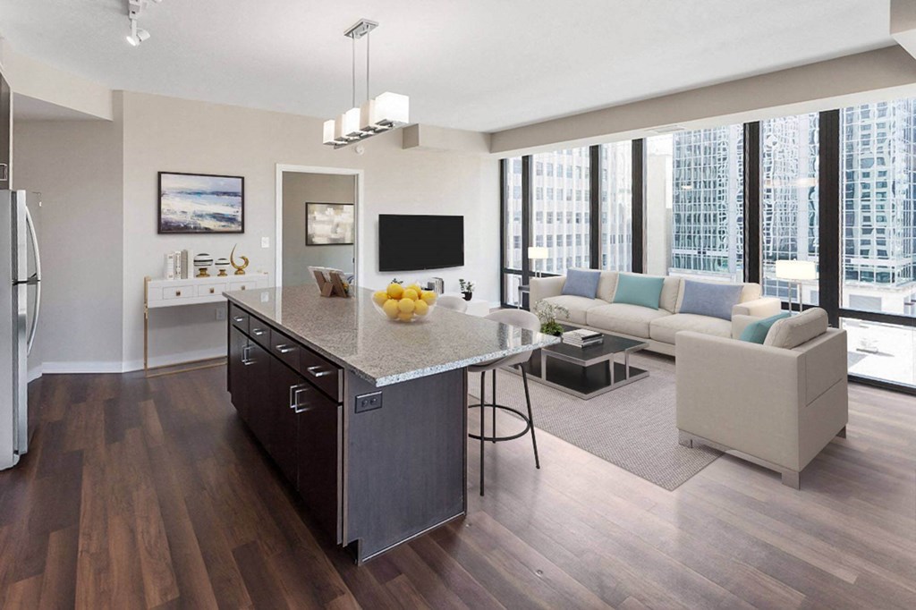 Modern open-plan living area here at Nic on 5th Apartment Homes with large windows offering a city view. Features a spacious kitchen island, cozy beige L-shaped sofa, and contemporary decor.