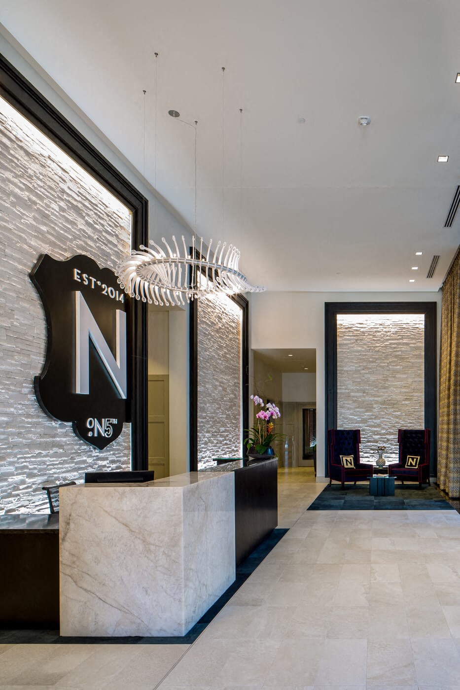 Modern lobby here at The Nic on 5th Apartment Homes with marble desk, geometric chandelier, and white textured walls. Two elegant chairs with cushions beside potted orchids create a welcoming ambiance.