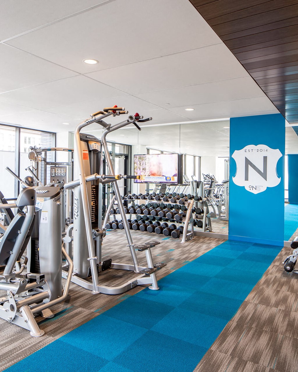 Modern gym here at Nic on 5th Apartment Homes with weight machines, mirrored wall, and a rack of dumbbells. A bold blue stripe runs across the floor, creating a vibrant, energizing atmosphere.