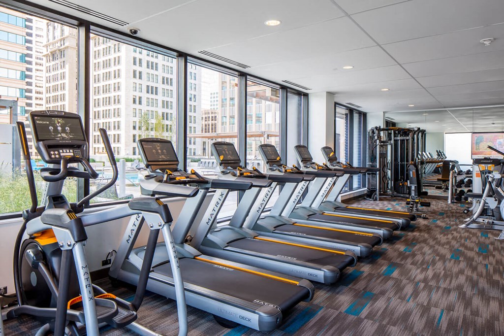 Modern gym interior here at Nic on 5th Apartment Homes with rows of treadmills and exercise equipment by large windows overlooking city buildings. Bright, clean, and spacious ambiance.