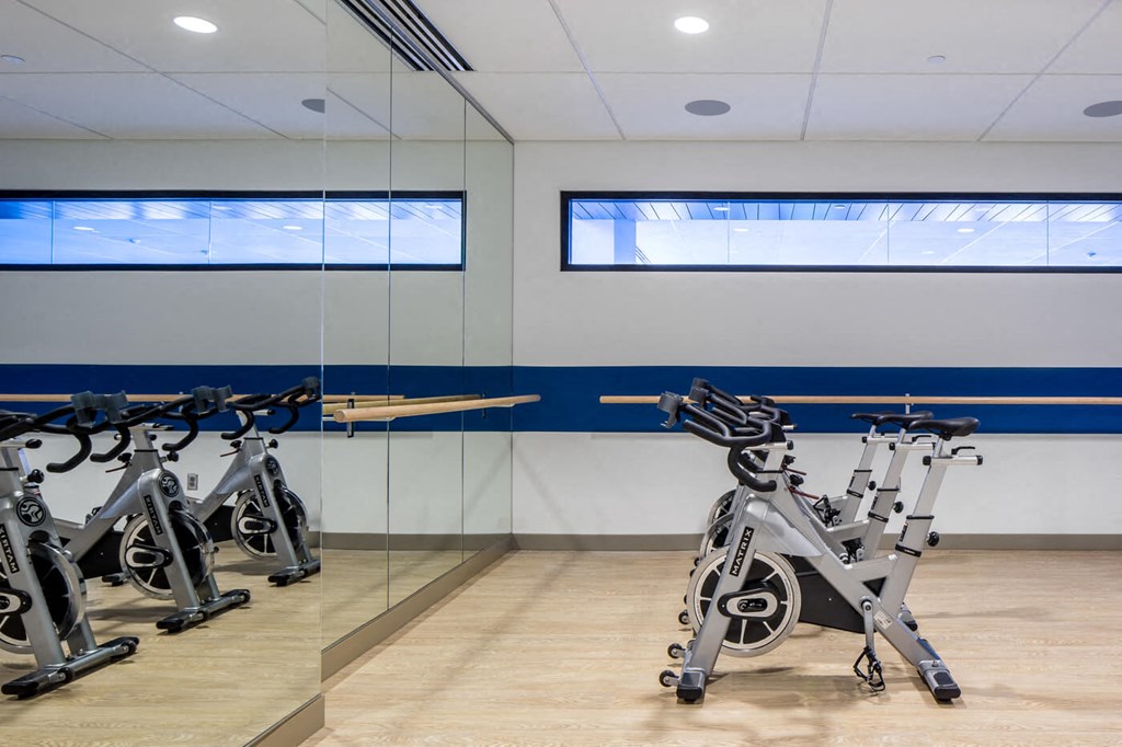 Modern gym room here at Nic on 5th Apartment Homes with sleek spin bikes lined up on polished wood floors. A mirrored wall reflects the bikes, creating a spacious, energetic atmosphere.