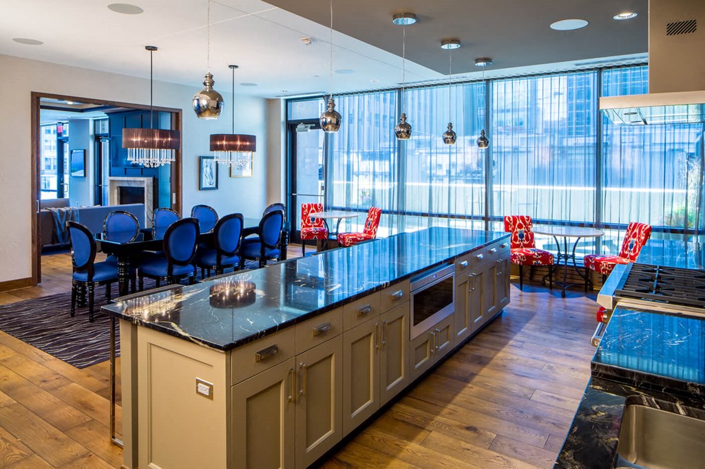 Modern kitchen and dining area here at Nic on 5th Apartment Homes with a sleek, black marble island, navy blue chairs around a dining table, and red patterned armchairs by large windows.