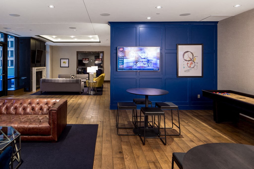 Modern lounge here at Nic on 5th Apartment Homes, with a brown leather sofa, gray chairs, blue accent wall featuring framed art and a TV, round table with stools, wood flooring.
