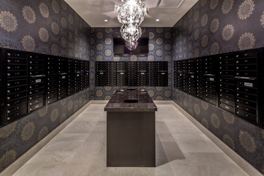 Luxurious mailroom, here at Nic on 5th Apartment Homes, with dark patterned wallpaper, glowing modern chandelier, and rows of black mailboxes. Central marble table adds elegance.