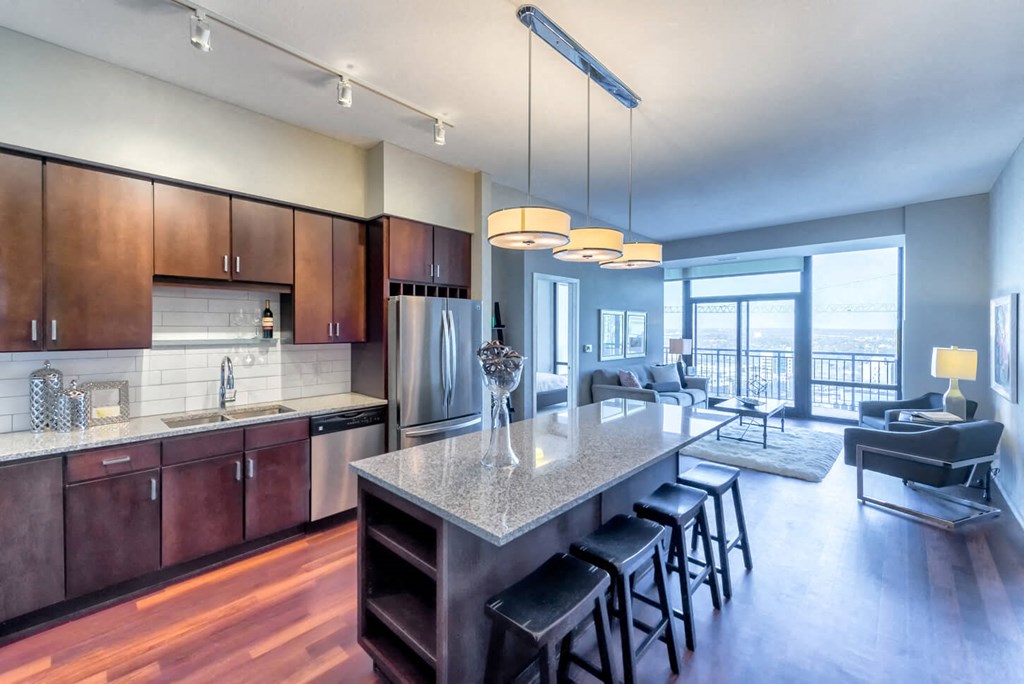 Modern kitchen and living area here at Nic on 5th Apartment Homes with wood flooring. Granite island with stools, stainless steel appliances, and pendant lights. Bright, open, and elegant.