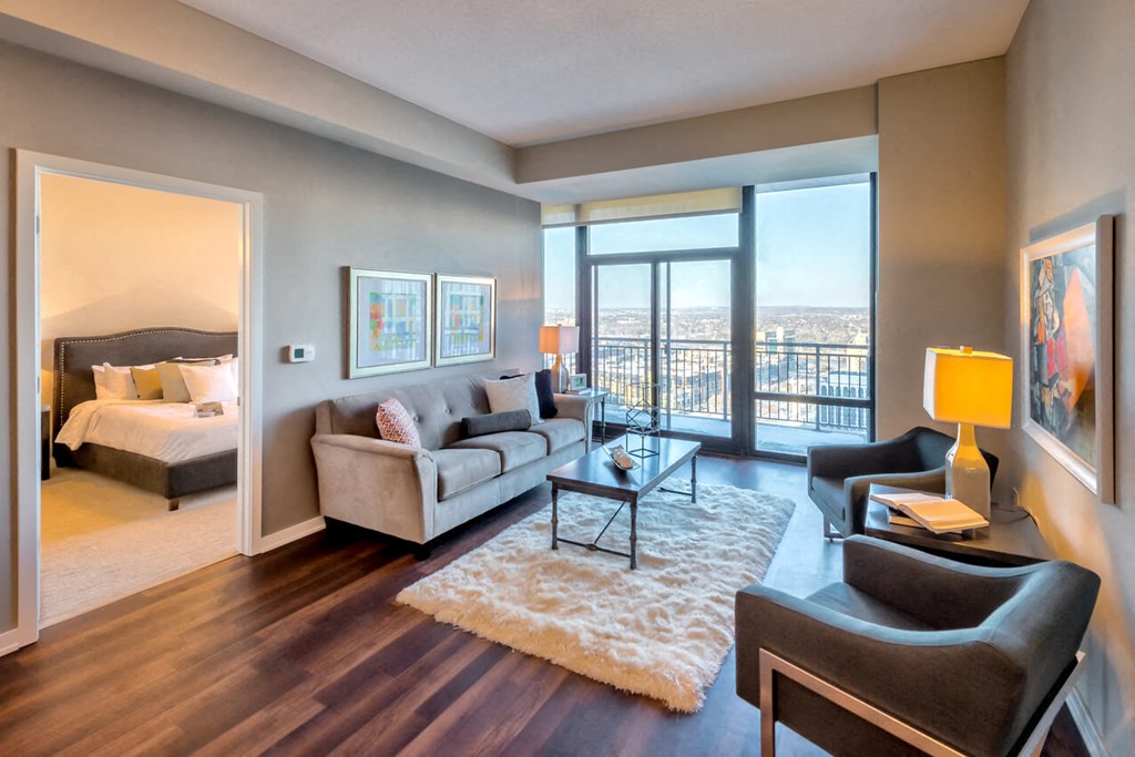 Modern living room here at Nic on 5th Apartment Homes with a cozy gray sofa, two armchairs, and a fluffy white rug. Large windows show a city view. Adjacent bedroom with neatly made bed.
