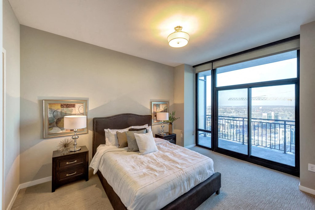 Modern bedroom here at Nic on 5th Apartment Homes with a cozy double bed, soft lighting from a ceiling fixture, framed art, and a balcony view through floor-to-ceiling windows. Calm ambiance.