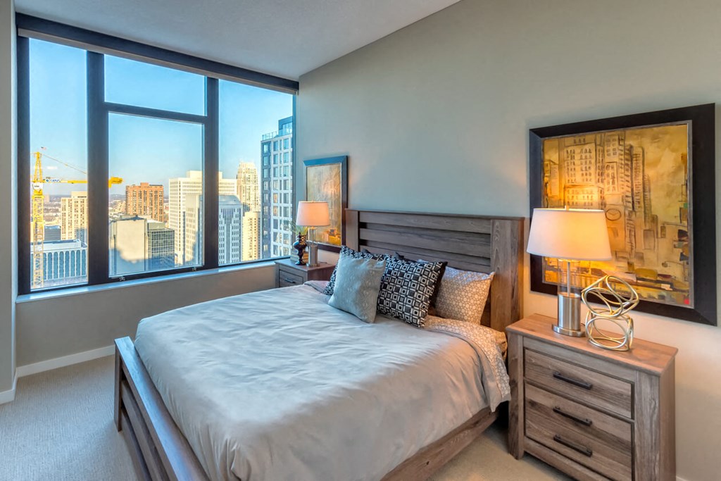 Modern bedroom here at Nic on 5th Apartment Homes with a wooden bed, patterned pillows, and two nightstands with lamps. Large window showcases a sunny cityscape view. Cozy and bright ambiance.