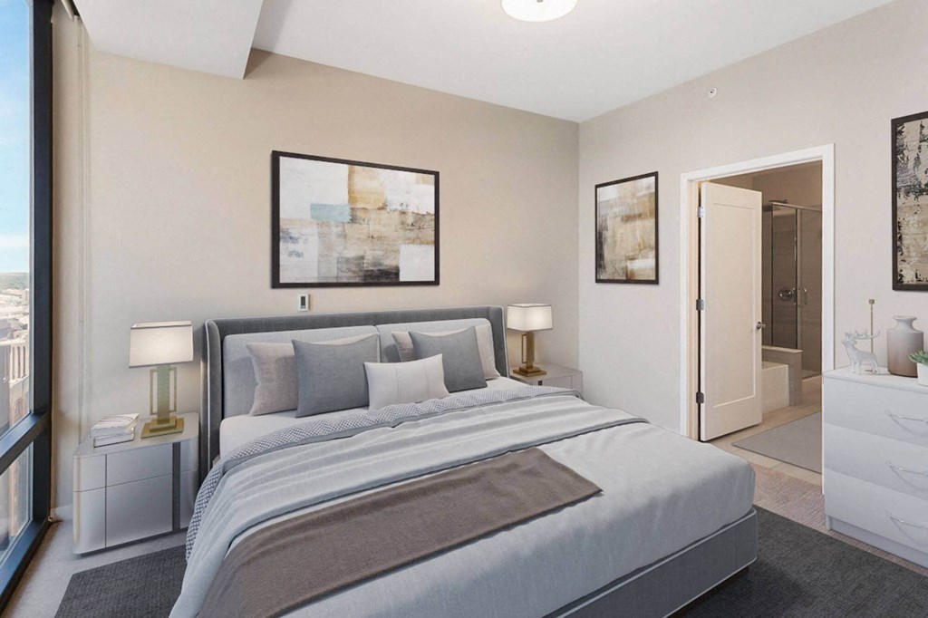 Modern bedroom here at Nic on 5th Apartment Homes with neutral tones, featuring a large bed with gray bedding, abstract art, bedside tables with lamps, and an open door showing a bathroom.