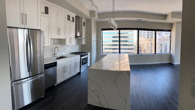 Modern kitchen, here at Nic on 5th Apartment Homes, with sleek white cabinets, marble countertops, and stainless steel appliances. Large windows offer a cityscape view, creating a bright, open atmosphere.