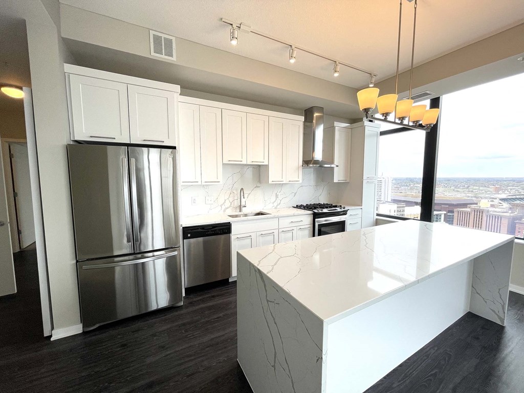 Modern kitchen here at Nic on 5th Apartment Homes with white cabinets, stainless steel appliances, and a large marbled island. A window offers a cityscape view, creating an airy feel.
