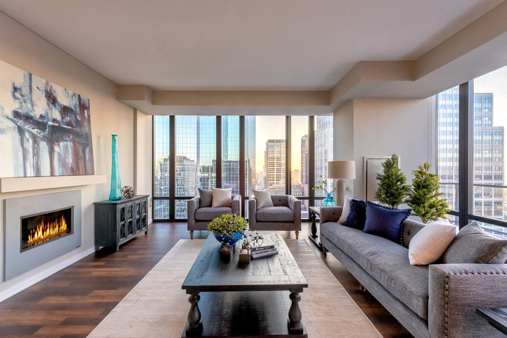 Modern living room here at Nic on 5th Apartment Homes with city view, large windows, gray sofas, a wooden coffee table, vibrant cushions, a lit fireplace, and abstract art. Cozy and elegant atmosphere.