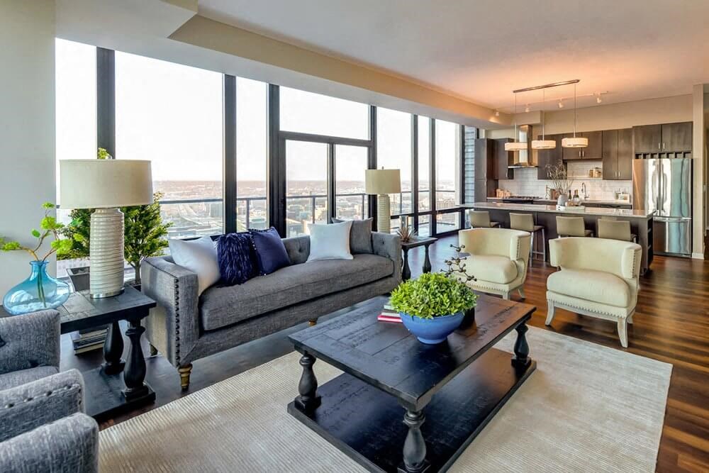 Modern living room here at Nic on 5th Apartment Homes with large windows showcasing a city view. Gray sofa, beige armchairs, dark coffee table with plants, and a sleek kitchen in the background.